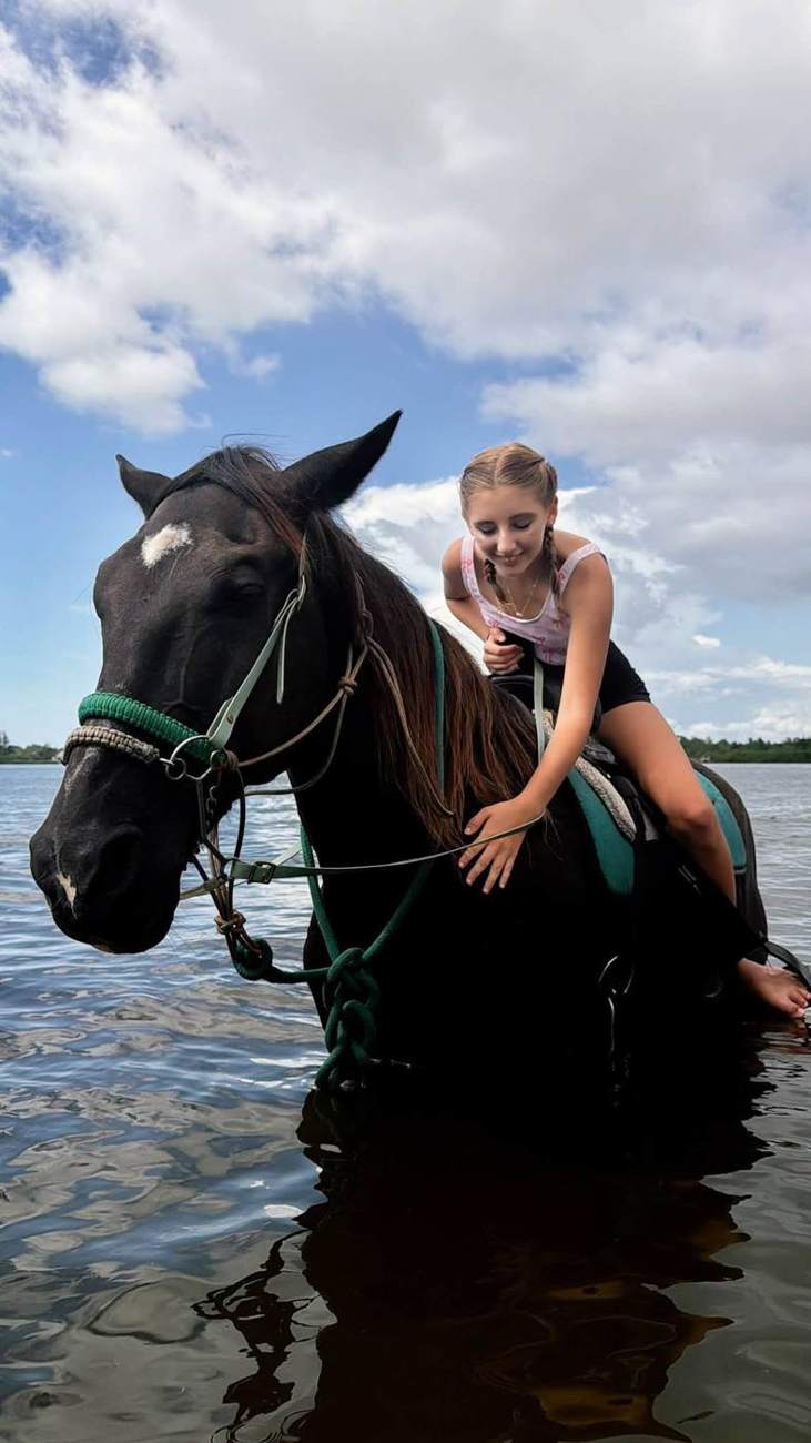 Kendra's daughter leaning down to pet a dark horse standing in shallow water