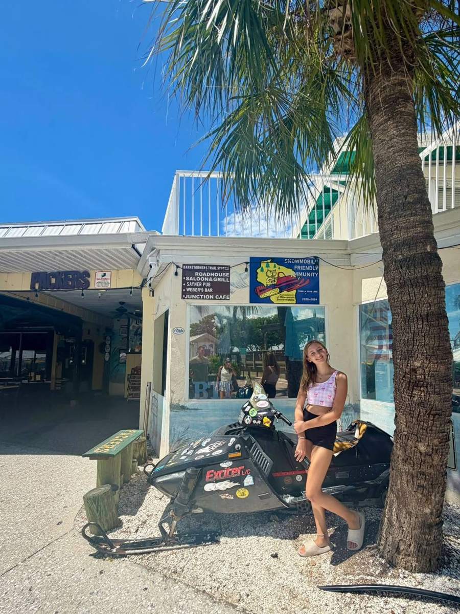 Kendra's daughter posing with a sticker-covered snowmobile under a palm tree outside the Packers bar
